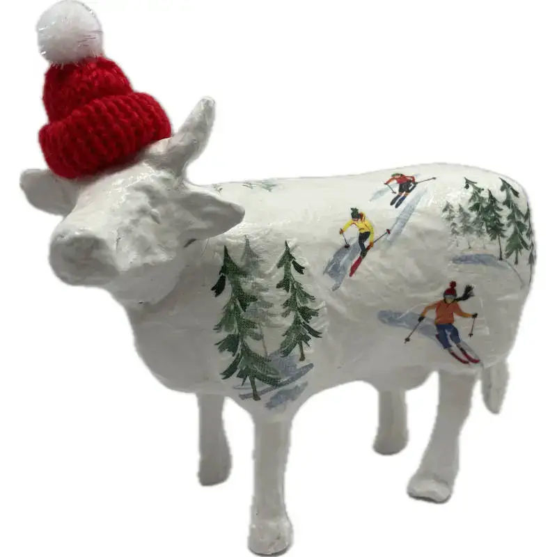 Vache décorative blanche en céramique ou résine, motif ski et paysages enneigés, portant un bonnet rouge avec pompon.