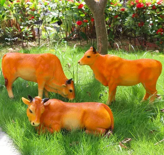 Trois vaches décoratives orange et jaune en peluche ou résine, posées dans une pelouse verdoyante avec arbustes colorés en arrière-plan.