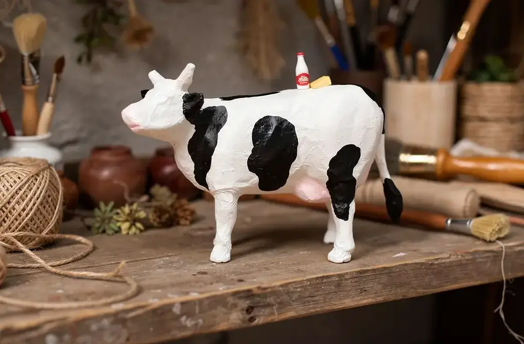 Figurine de vache artisanale en papier mâché posée sur une étagère en bois dans un atelier artisanal.